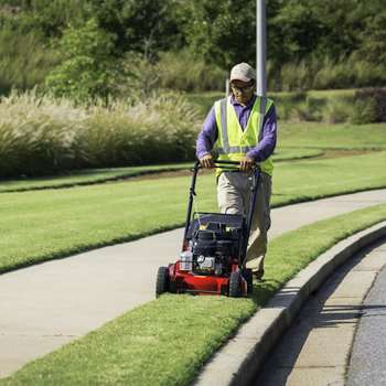 Landscaper mowing grass with an Exmark Commercial 21 X-Series walk-behind mower along a street.
